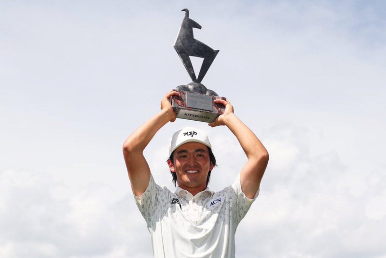 Smiling man in a white polo and cap raises a trophy above his head against a blue sky.