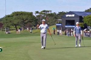 Two golfers walk on a sunny golf course during a tournament, with spectators and a large sponsor tent in the background.