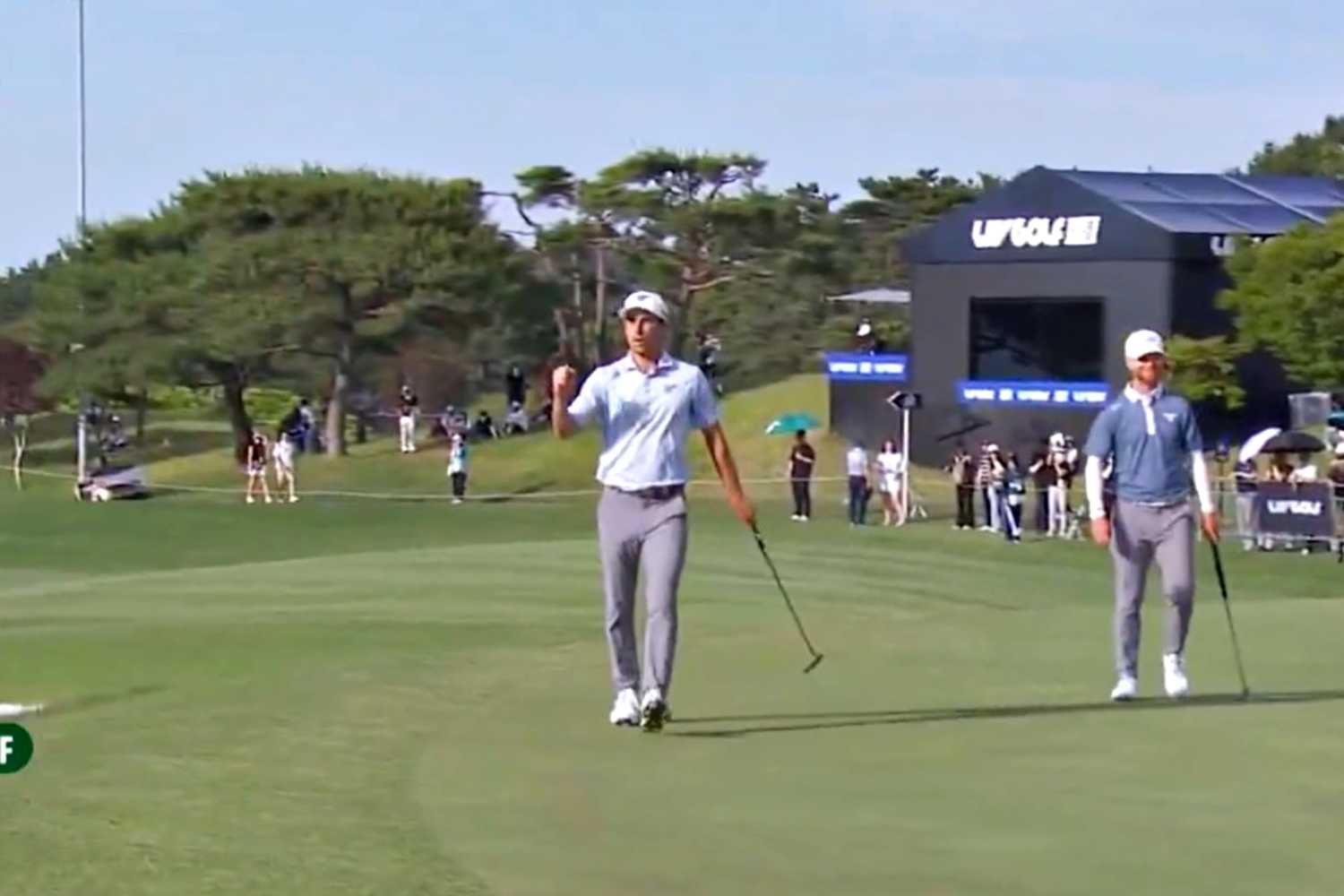 Two golfers walk on a sunny golf course during a tournament, with spectators and a large sponsor tent in the background.