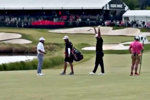 Four golfers on a golf course fairway near sand bunkers; the lead player raises a club overhead while others walk with bags in tow.