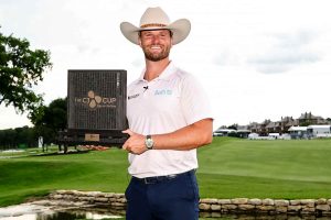 Man wearing a cowboy hat and polo shirt holds a trophy plaque on a golf course, smiling at the camera. The CJ Cup Byron Nelson trophy is visible.