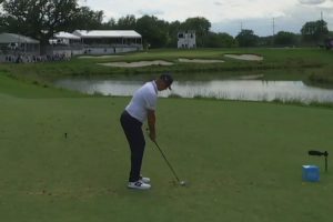 Golfer in a white polo and cap prepares to swing from the tee on a green course, with a water hazard and stands in the background.