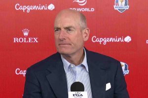Man in a dark suit and striped shirt speaks into a PGA microphone at a press conference with a red backdrop featuring Capgemini and Rolex logos.