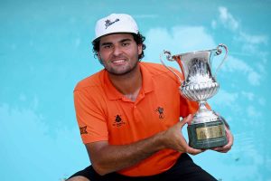 Man in an orange polo shirt and white cap holds a large silver trophy beside a blue pool, smiling at the camera.