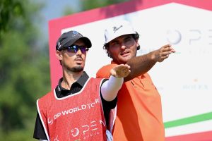 Two golfers wearing red bibs, the man in a black cap and sunglasses pointing to the side while the other in a white cap extends his arm, during a tournament backdrop.