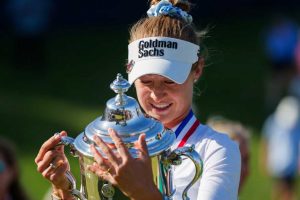 Female golfer in a white visor with Goldman Sachs logo, holding a shiny trophy and smiling in celebration.