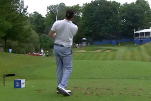 Golfer in white shirt and light blue pants swinging a club from the tee on a lush golf course.