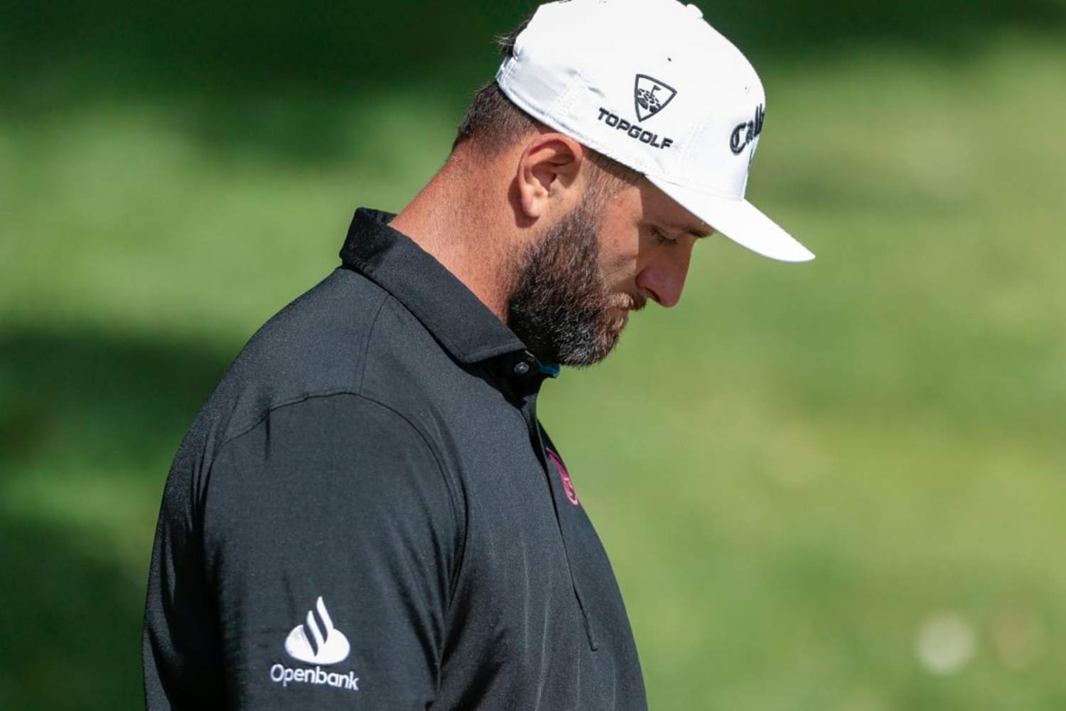 Man in a black polo shirt and white golf cap looking down, on a grassy outdoor course; Openbank logo on sleeve.