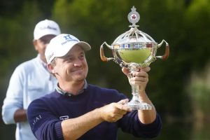 Man in a navy sweater holding up a shiny trophy with a smile, outdoors during daytime prize ceremony