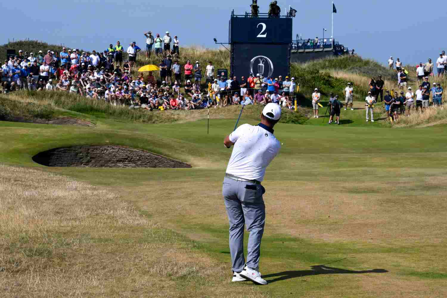 Golfer in a white shirt swinging on a sunny golf course while a large crowd watches from the stands and rough ground nearby.