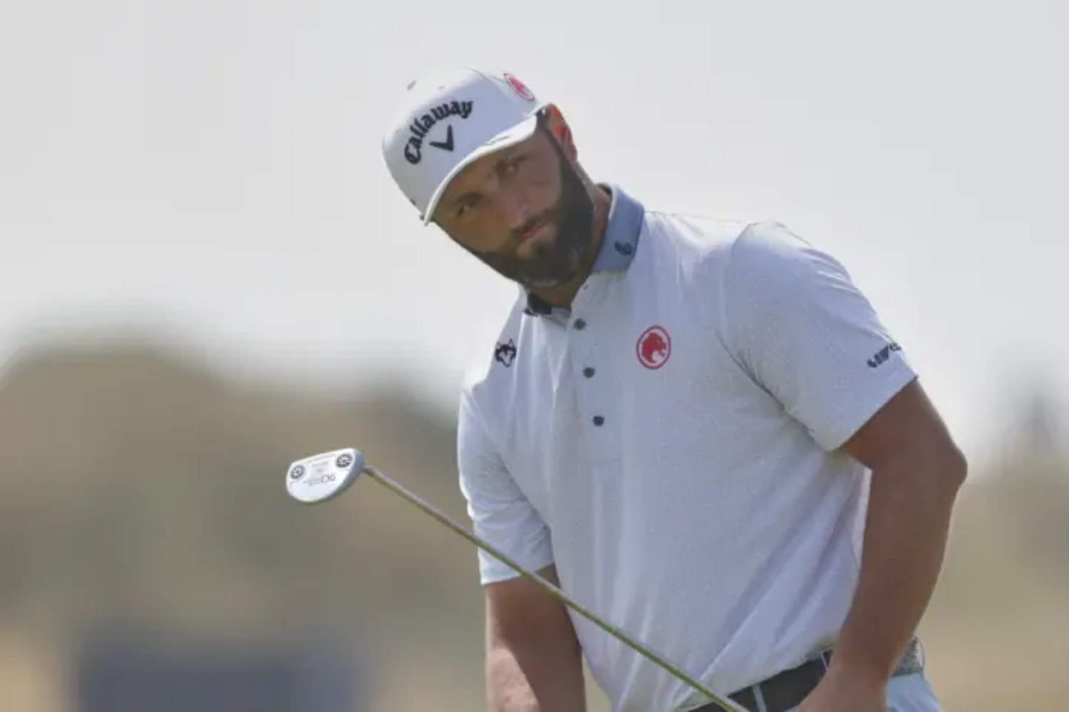 Male golfer with a beard in a white polo and Callaway cap preparing to swing with a club on a sunny day.
