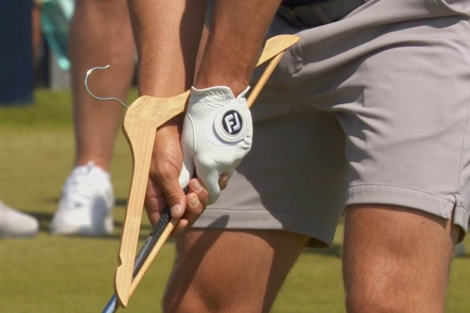 Close-up of a golfer's gloved hand gripping a wooden golf club on the grass outside the tee area with a visible FJ logo on the glove.