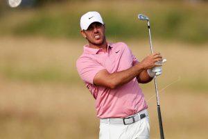 Male golfer in pink polo and white cap swings a golf club on a course, mid-backswing with a focused expression.