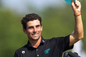 Smiling man outdoors, wearing a black polo, raising his right arm in victory beside a microphone stand with a teal logo on his shirt