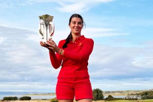Young woman in red athletic outfit holds a silver trophy outdoors against a blue sky.